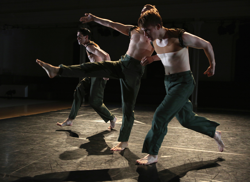 three shirtless male dancers perform on a dimly lit stage